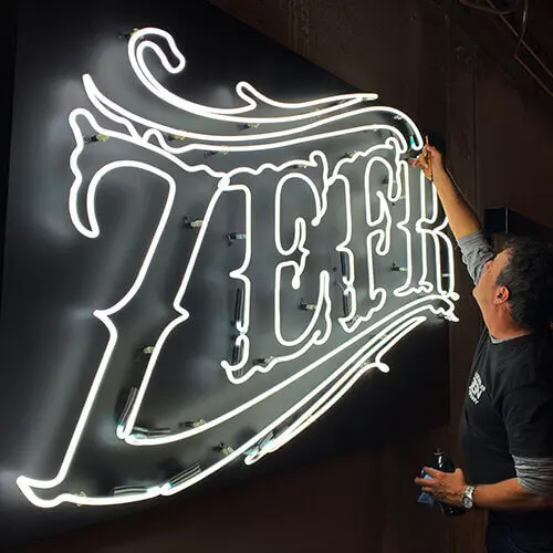 Skilled craftsman fine-tuning illuminated neon sign 'ZEPH' with elegant script against dark background, showcasing precision and artistry in custom neon signage.